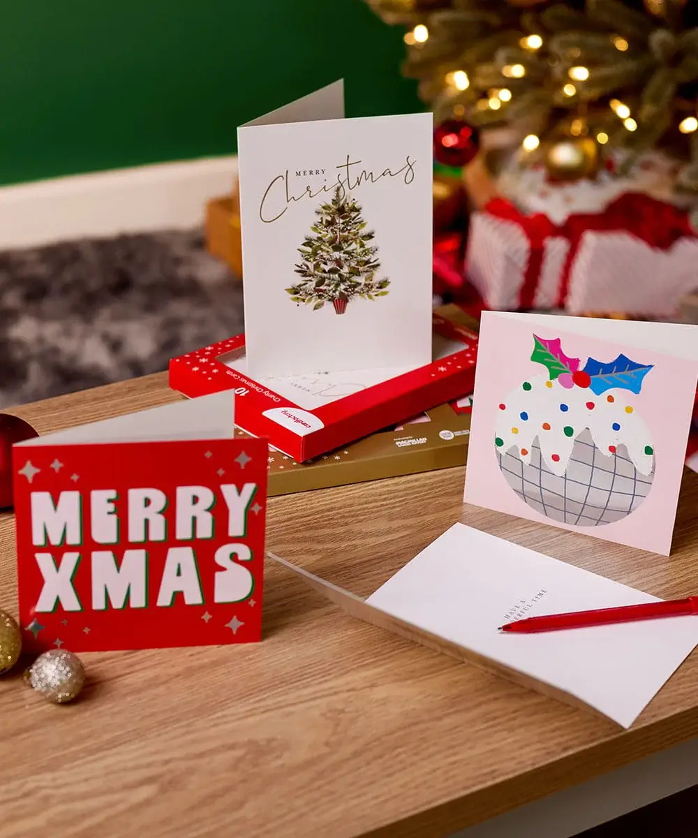 Selection of Christmas greeting cards displayed on a wooden table with festive decorations, gift boxes, baubles, and a decorated Christmas tree in the background. Selection of Christmas greeting cards displayed on a wooden table with festive decorations, gift boxes, baubles, and a decorated Christmas tree in the background.
